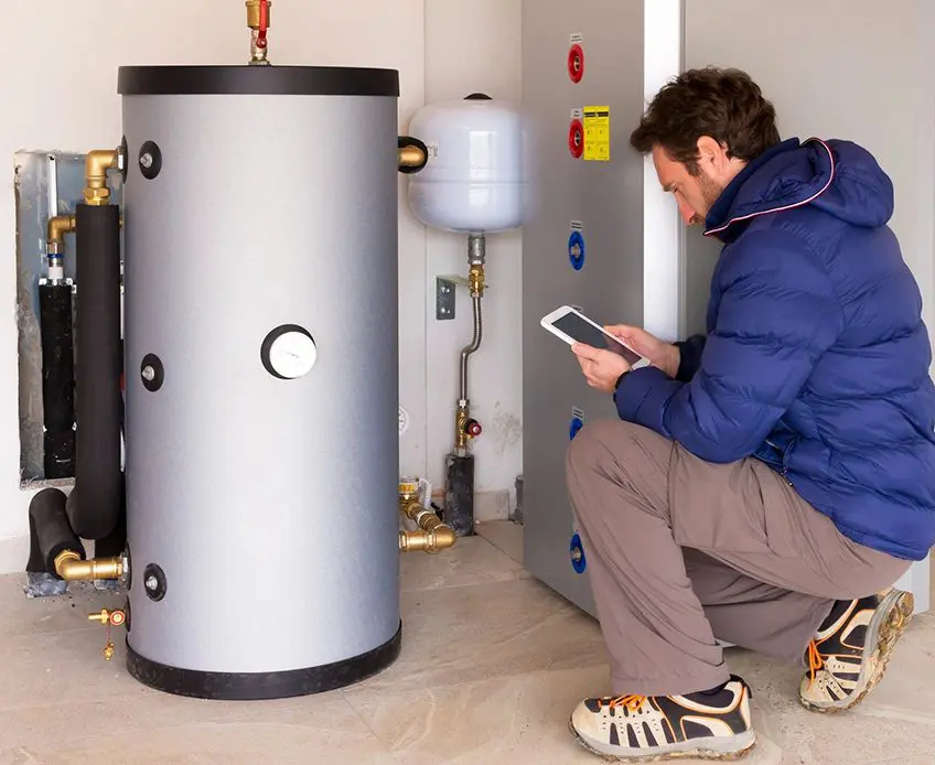 A man is looking at his tablet in front of an electric water heater.